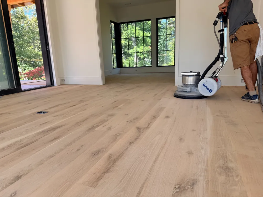 Person sanding and refinishing a wood floor in a bright room with large windows and natural light.