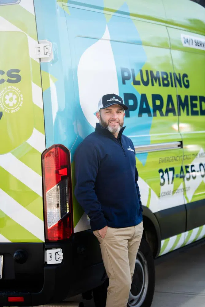 Man in navy blue jacket and cap stands beside a green and blue Plumbing Paramedics service van.