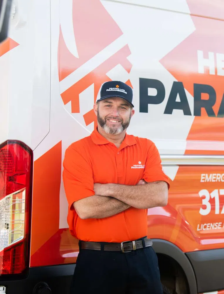 Smiling technician in orange uniform and cap standing with arms crossed beside branded paramedic service van.