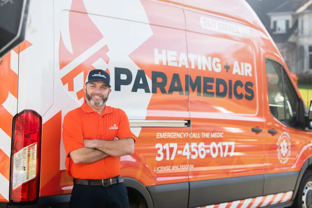Man in orange uniform stands confidently next to a service van for Heating + Air Paramedics.