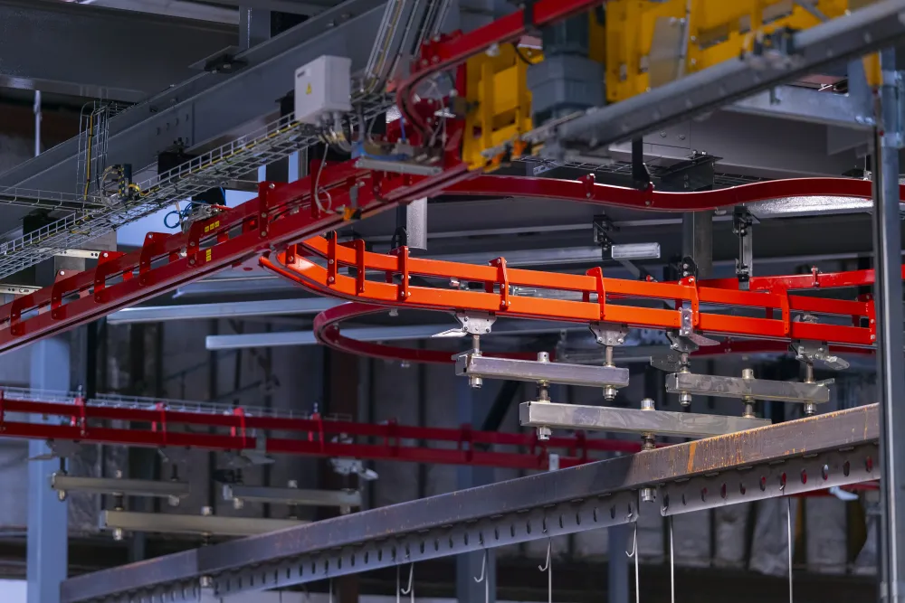 Industrial conveyor system with red and orange rails moving through a manufacturing facility ceiling.