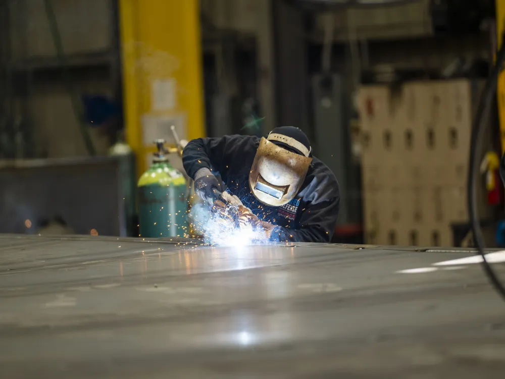 Welder wearing protective gear sparks bright light welding metal sheet in industrial workshop.