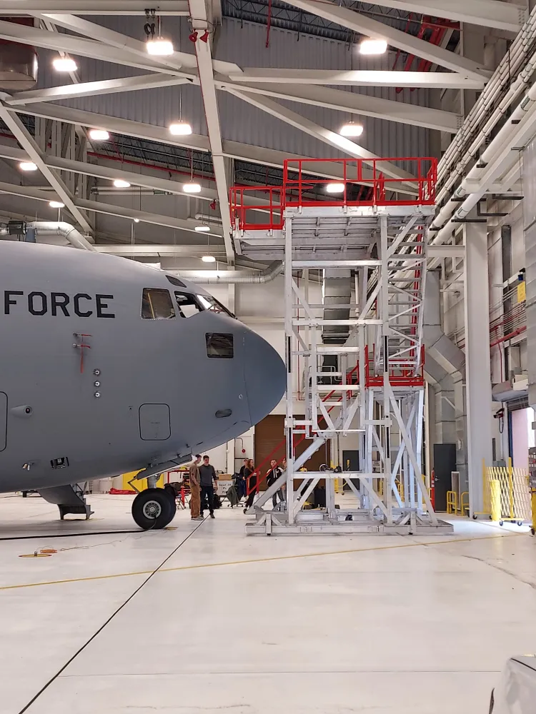 Military aircraft inside a hangar with a maintenance platform and personnel working nearby.