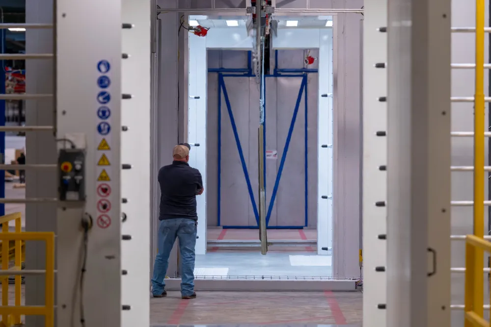 Man in a cap observing a large industrial machine in a spacious factory setting with white and blue structural elements.