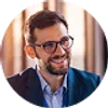 Smiling man with glasses and beard wearing a suit jacket in a warmly lit indoor setting