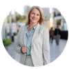 Smiling middle-aged woman in light gray suit posing outdoors with a blurred urban background.