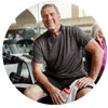 Smiling middle-aged man in workout clothes holding water bottle at gym with equipment in background.