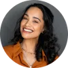 Smiling woman with curly dark hair wearing an orange shirt against a gray background