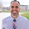Smiling man wearing a light purple shirt and colorful plaid tie outdoors with blurred buildings and grass background