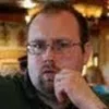 Close-up of a man with glasses resting his chin on his hand in a warmly lit indoor setting