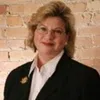 Professional woman with glasses and short blonde hair wearing black blazer and white shirt against brick wall.