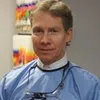 Male dentist wearing a blue dental gown and magnifying glasses standing in a clinic with colorful charts in background