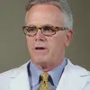 Mature male doctor wearing glasses, white coat, yellow tie, speaking in a professional setting with plant background