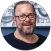 Smiling middle-aged man with glasses and a beard in a black shirt, with abstract art in the background.