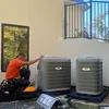 Estes Services Technician in orange shirt installing three Carrier outdoor air conditioning units beside a modern house on sunny day