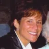 Smiling woman with short brown hair wearing a black jacket and white shirt in a dimly lit setting.