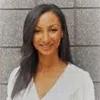 Smiling woman with long dark hair, wearing a white blouse against a gray brick background.