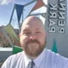 Bearded man in white shirt and tie smiling outdoors with colorful geometric sculpture and sign in background
