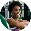 Smiling woman with curly hair in purple top resting on gym equipment in fitness center.