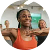 Smiling woman in orange tank top exercising with group in bright gym environment.