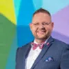 Smiling man in a blue suit and pink bow tie stands before a colorful geometric wall mural.