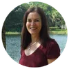 Smiling woman with long brown hair wearing a maroon top standing outdoors near a lake with trees in background