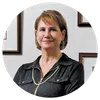 Middle-aged woman with short light brown hair wearing a dark blouse, smiling against a white wall with framed pictures.