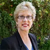 Smiling woman with short blonde hair, glasses, red lipstick, black blazer, and blue top, standing outdoors with greenery.