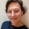 Close-up portrait of a smiling woman with short hair against a light textured wall, wearing a dark top.