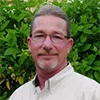 Middle-aged man with glasses and gray hair standing in front of green leafy plants wearing a white shirt