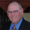 Smiling elderly man wearing glasses, a blue shirt, striped tie, and dark suit jacket in an indoor setting.
