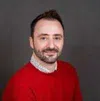Smiling man with short dark hair and beard wearing a red sweater over a patterned shirt on gray background