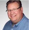 Smiling man wearing glasses and a blue checkered shirt against a plain light background.