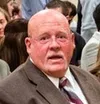 Elderly man in a brown suit and striped tie in a crowded indoor setting with people in the background
