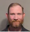 Close-up of a man with a red beard, short hair, and blue eyes against a plain gray background.