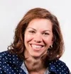 Smiling woman with brown hair wearing a navy blue polka dot blouse against a white background