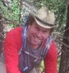 Smiling man wearing a cowboy hat and red jacket hiking in a forested area
