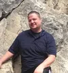 Man wearing a dark polo shirt sitting and smiling against a rocky background outdoors
