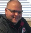 Smiling bald man with glasses wearing a dark jacket with a University of Utah logo inside a room with window blinds.