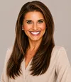 Smiling woman with long brown hair wearing pearl earrings and beige top against plain background