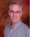 Middle-aged man with gray hair and glasses smiling against a warm brown backdrop, wearing a patterned button-up shirt