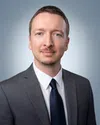 Professional portrait of a man in a gray suit, white shirt, and navy tie against a light blue background.