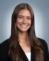 Smiling young woman with long brown hair wearing a black blazer and a cross necklace against a gray background.