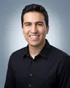Smiling young man with dark hair wearing a black button-up shirt against a light gray background