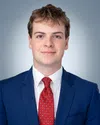 Young man in a blue suit and red patterned tie smiling with a neutral gray background.