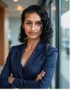Confident woman with curly hair wearing a navy blazer standing with arms crossed near a window.