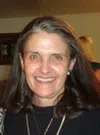 Smiling middle-aged woman with long brown hair wearing a black top and pearl earrings indoors.