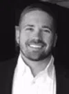Smiling man with short hair and beard wearing a white shirt and dark jacket against black background