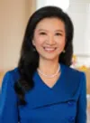 Smiling woman with long black hair wearing a blue dress and pearl necklace in an indoor setting