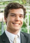 Young man smiling in a suit and tie with a green outdoor background and a white fence.